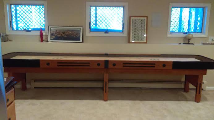 A Champion 9' The Grand Champion Shuffleboard Table sits beneath three windows in the room.