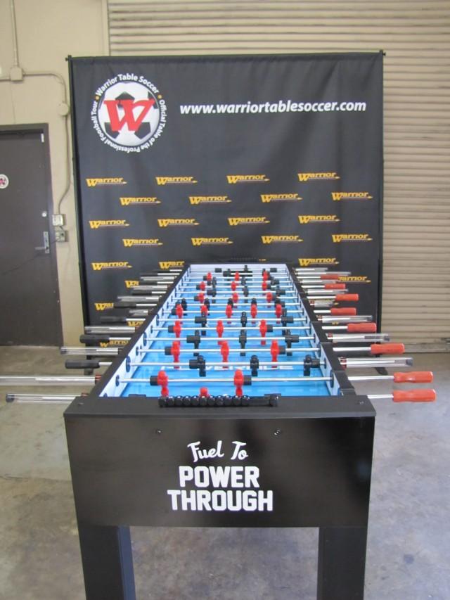 A Warrior 8 Players Foosball Table sits before a branded Warrior Table Soccer banner.
