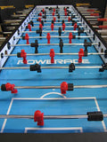 A close-up of the Warrior 8 Players Foosball Table with rod guard and red/black players on blue.